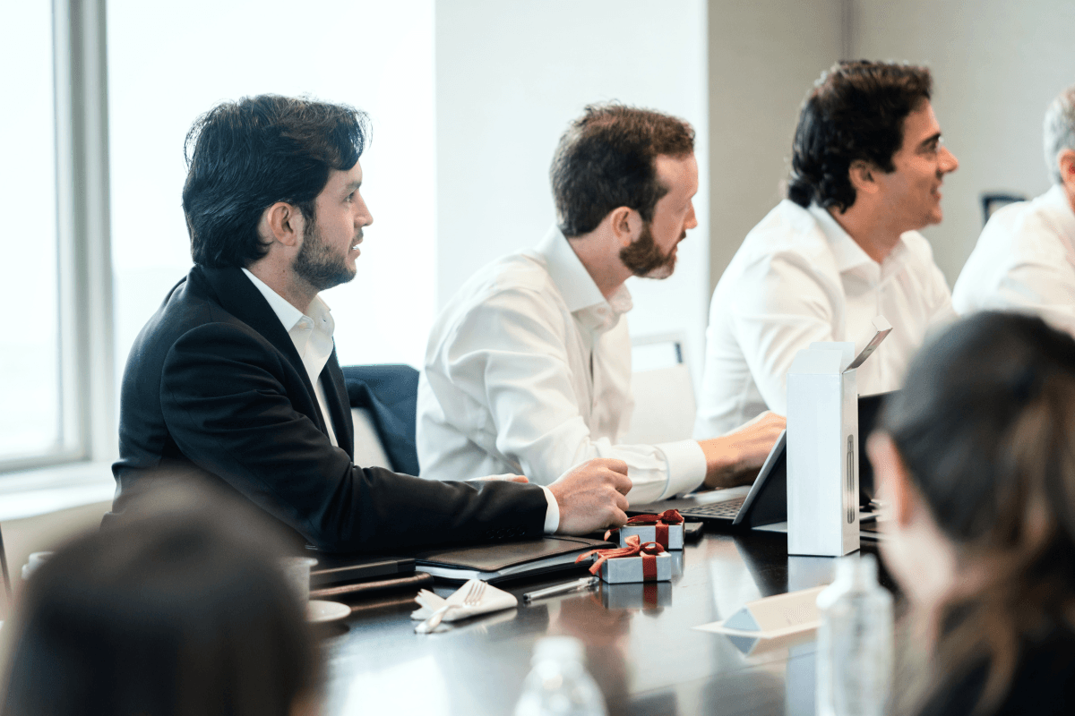 Three men in business attire sit at a conference table with laptops and papers, actively participating in a meeting.