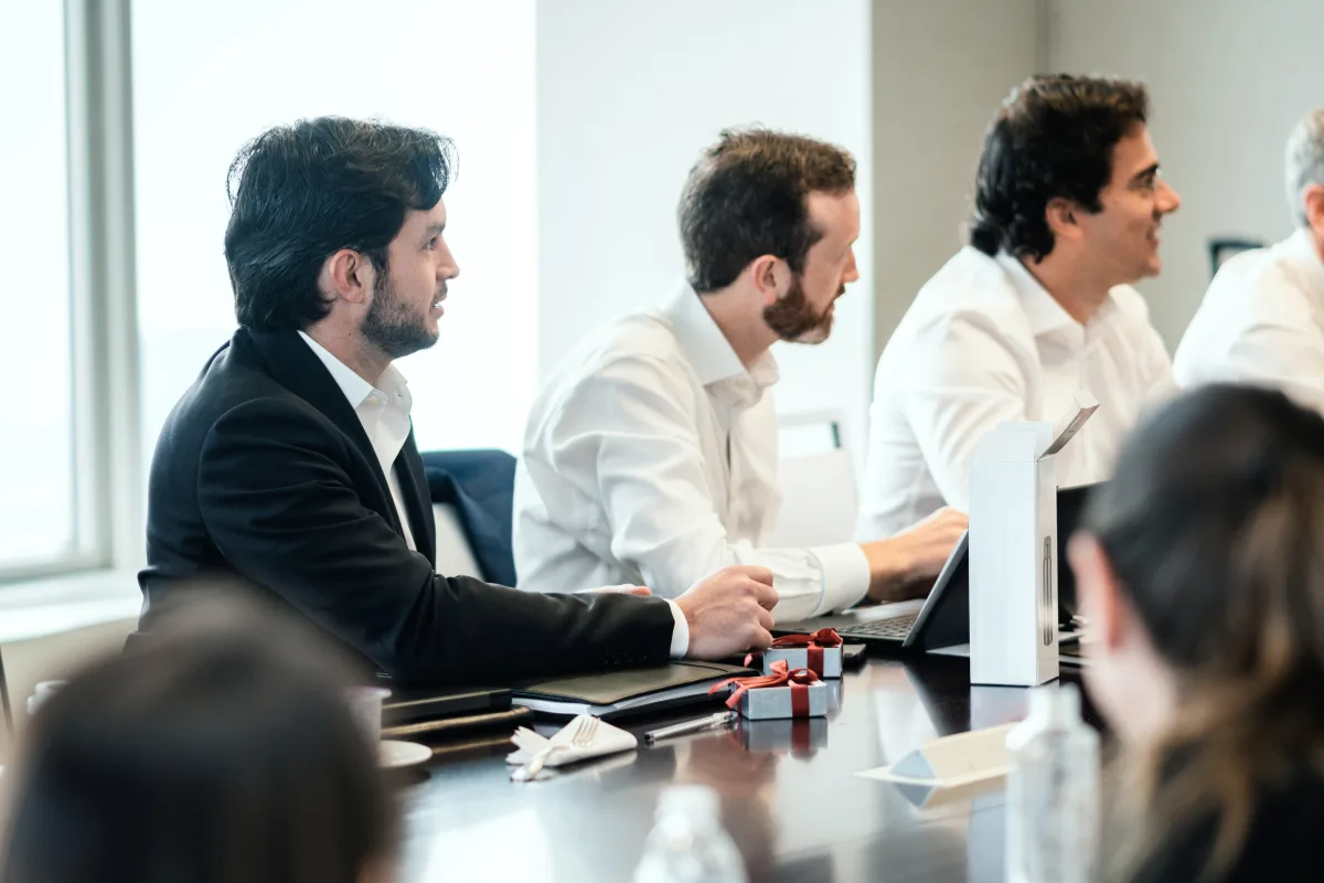 Three men in business attire sit at a conference table with laptops and papers, actively participating in a meeting.