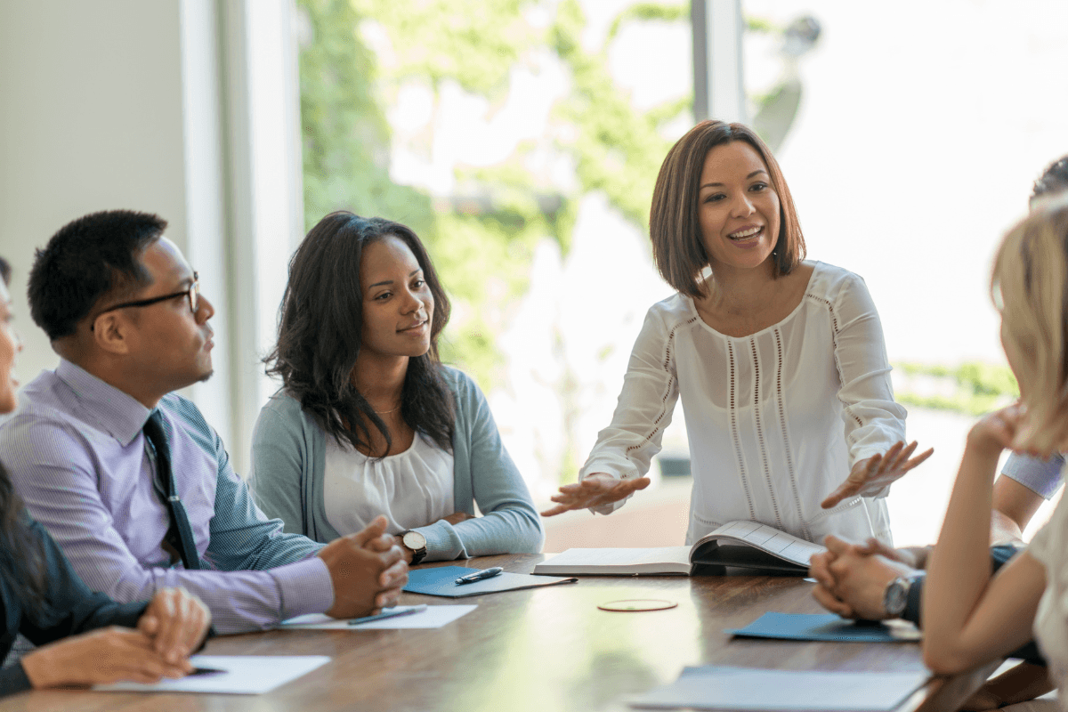 A group of people sit around a table in a meeting, with one woman speaking and gesturing while others listen attentively - Psychological Safety