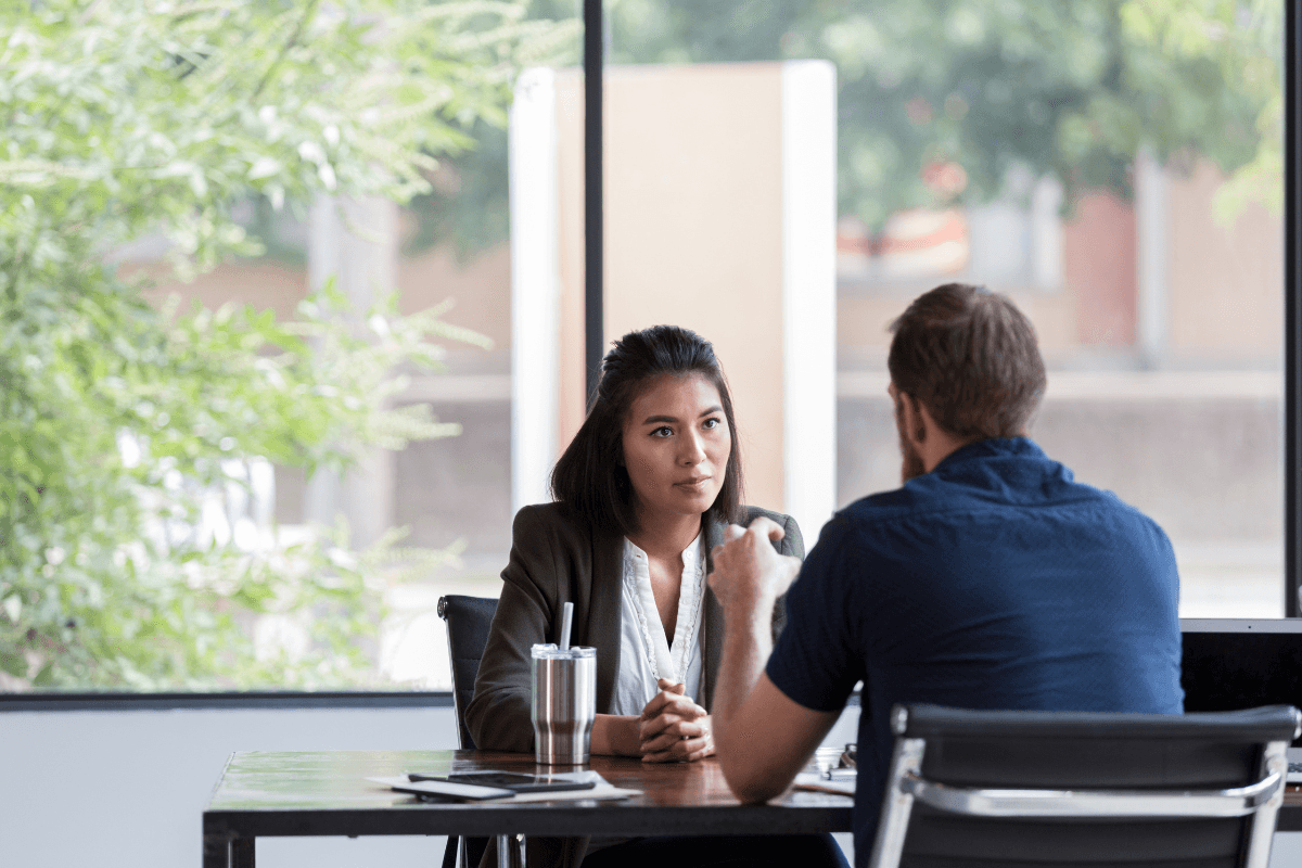 Two people sit across from each other at a table in an office, engaged in a serious conversation about team performance. Papers, a laptop, and a drink tumbler are on the table. Large window in background.
