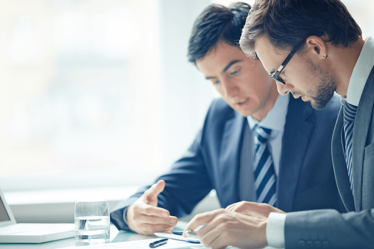 Two men in business suits sit at a desk discussing documents, with a glass of water, pen, and tablet visible—focusing on team effectiveness during their meeting.