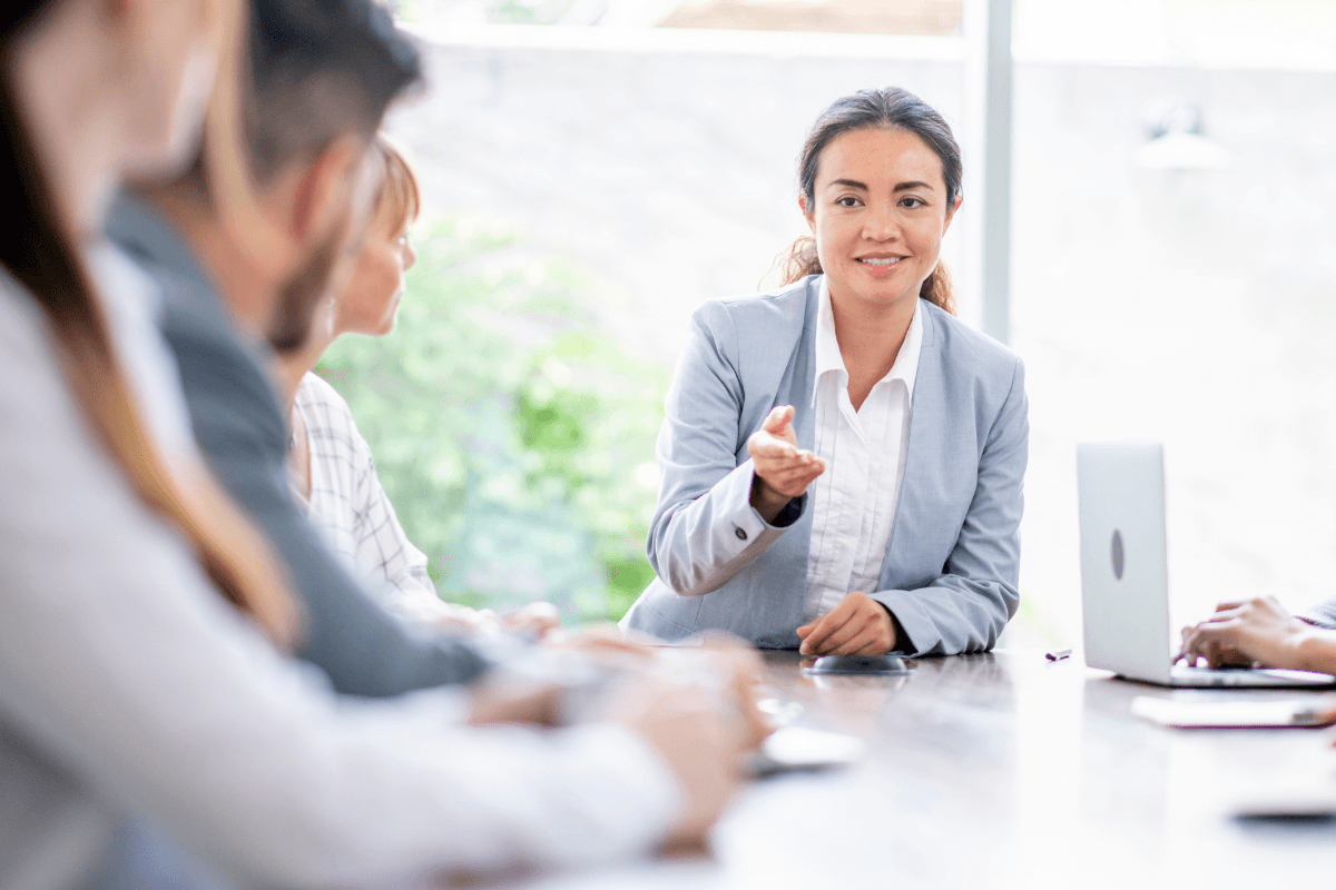 A woman in a light suit discusses team performance with colleagues at a conference table in a bright office, with a laptop and documents visible on the table.