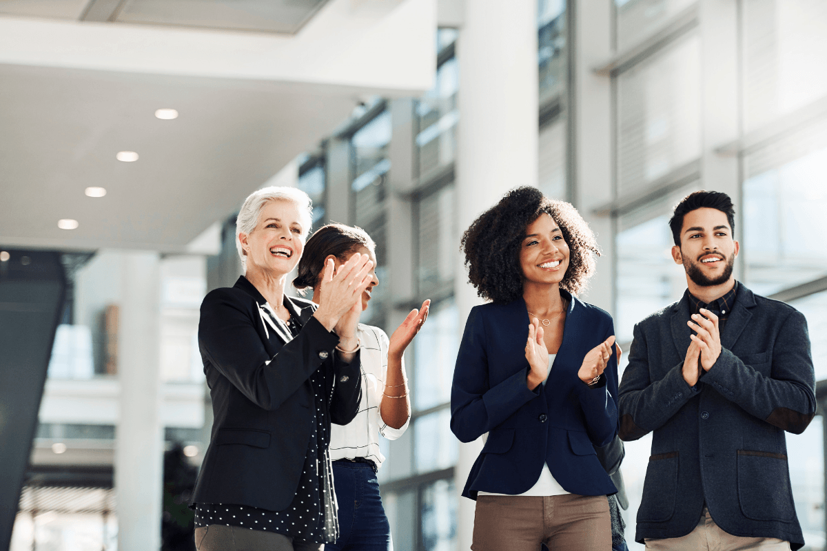 Four professionally dressed people stand indoors, smiling and clapping in a brightly lit, modern office space that fosters psychological safety and celebrates strong team performance.