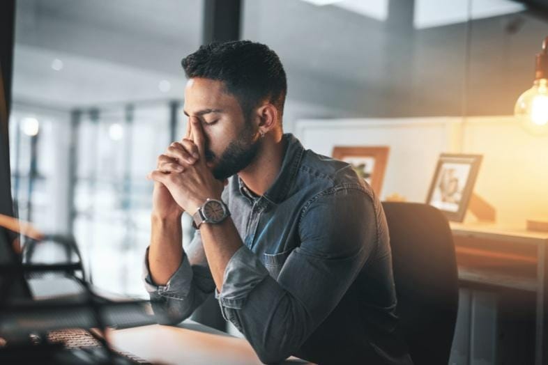 A man sits at a desk with his eyes closed and hands pressed to his face, appearing stressed or deep in thought in an office setting.