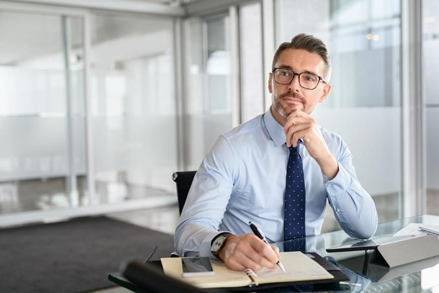 A man in business attire sits at a desk in an office, holding a pen and writing thoughtfully in a notebook, conveying a strong, focused presence at peak performance.