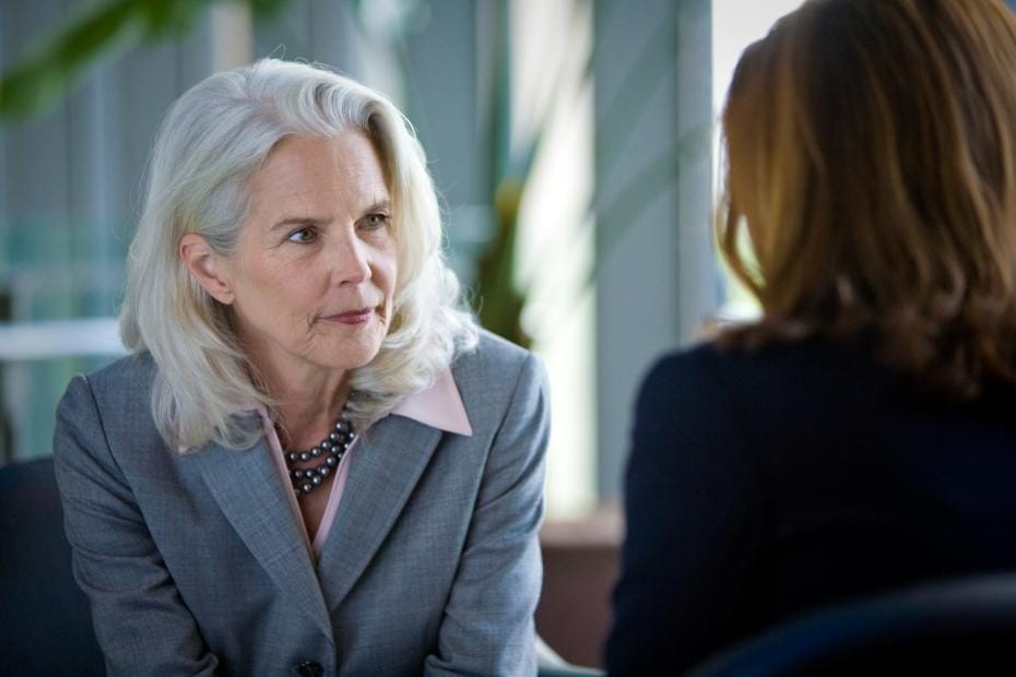 Two women in business attire sit facing each other in conversation; one has gray hair and is listening attentively.