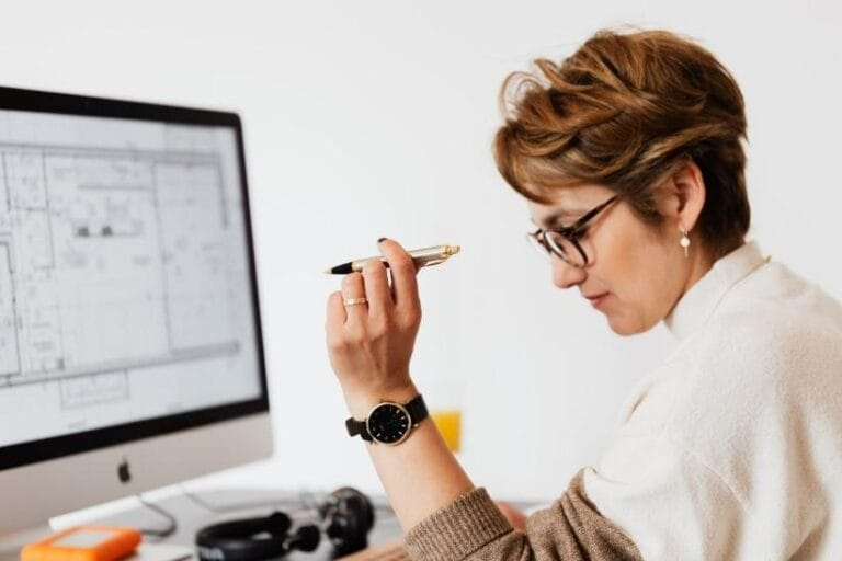 Person holding a pen and looking at architectural plans on a computer monitor at a desk.