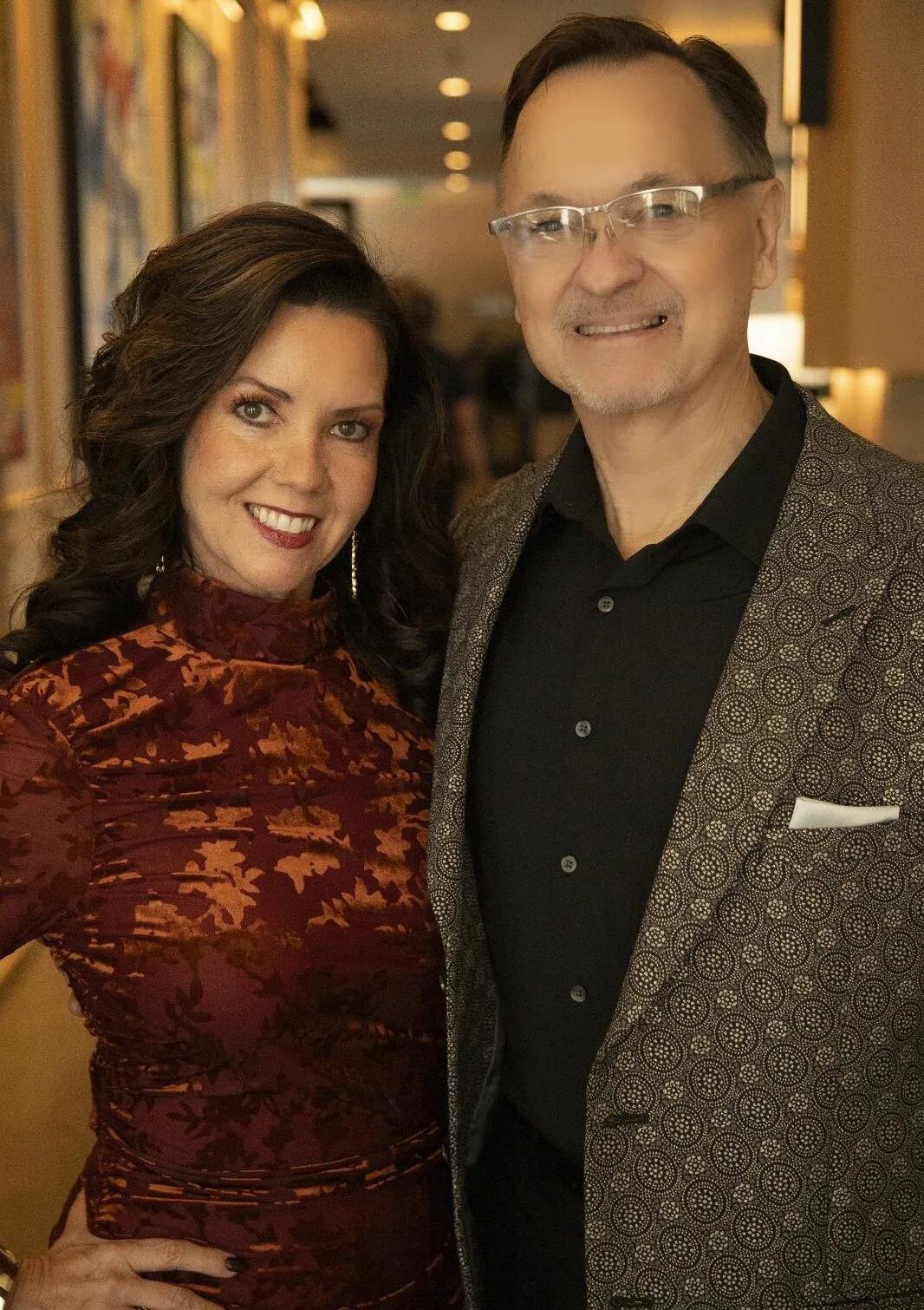 A woman in a patterned red dress and a man in a black shirt and patterned blazer, both with backgrounds in consulting, stand side by side indoors, smiling at the camera.