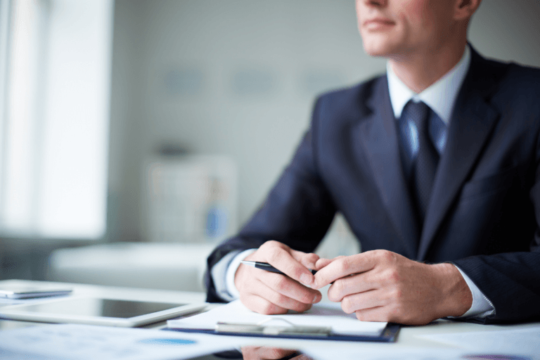 A man in a suit sits at a desk with documents, a tablet, and a pen in hand, exuding confidence as he engages in business conversations in an office setting.