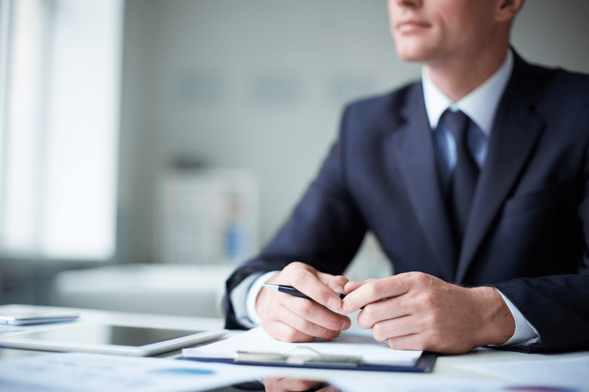 A man in a suit sits at a desk with documents, a tablet, and a pen in hand, exuding confidence as he engages in business conversations in an office setting.