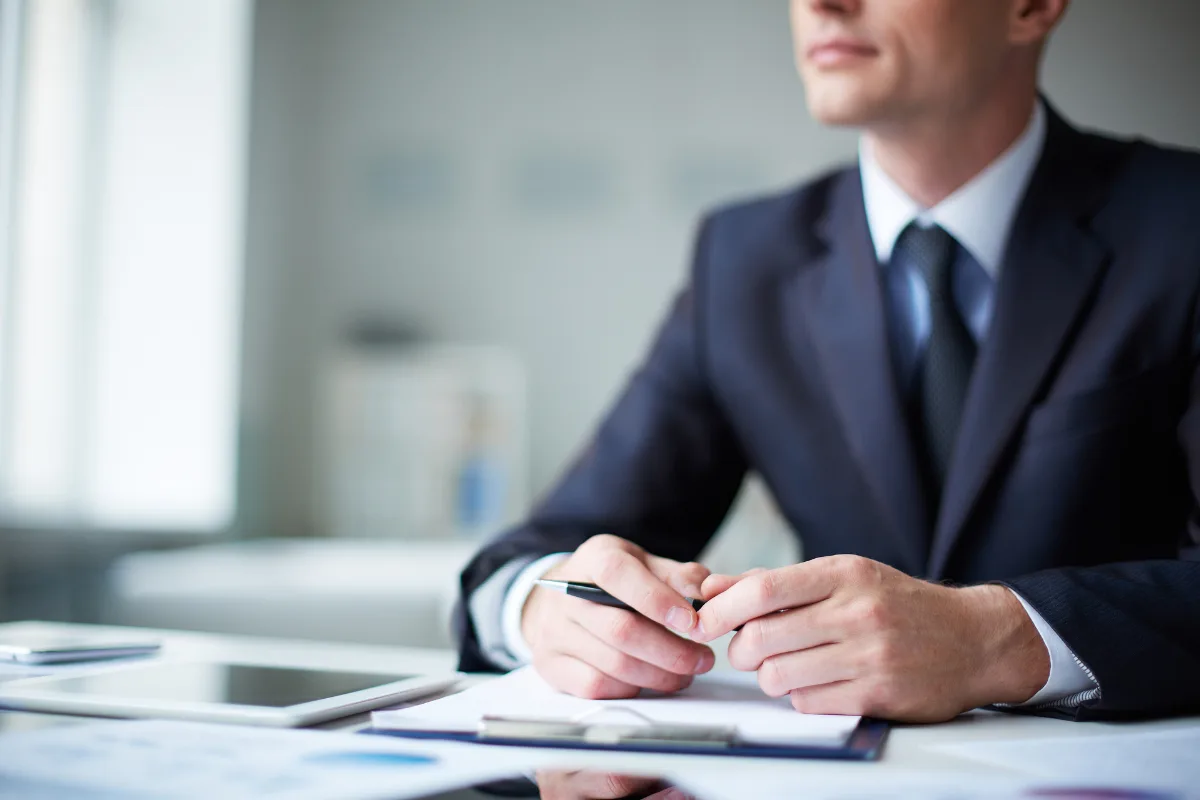 A man in a suit sits at a desk with documents, a tablet, and a pen in hand, exuding confidence as he engages in business conversations in an office setting.