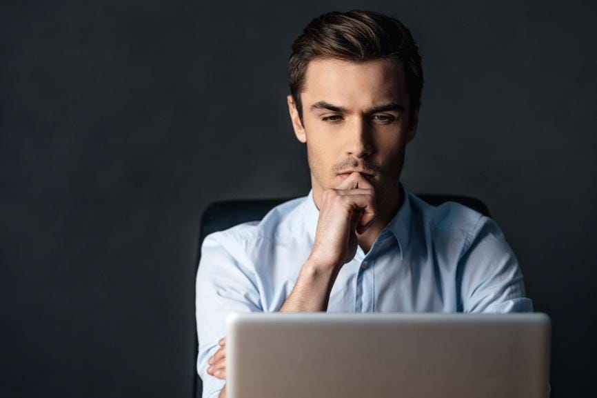 A man in a light blue shirt sits at a desk, looking intently at a laptop screen with his hand resting on his chin.