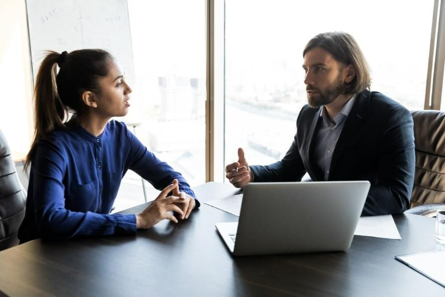 A woman and a man sit at a desk with a laptop, engaged in a serious Conversations in a modern office setting.