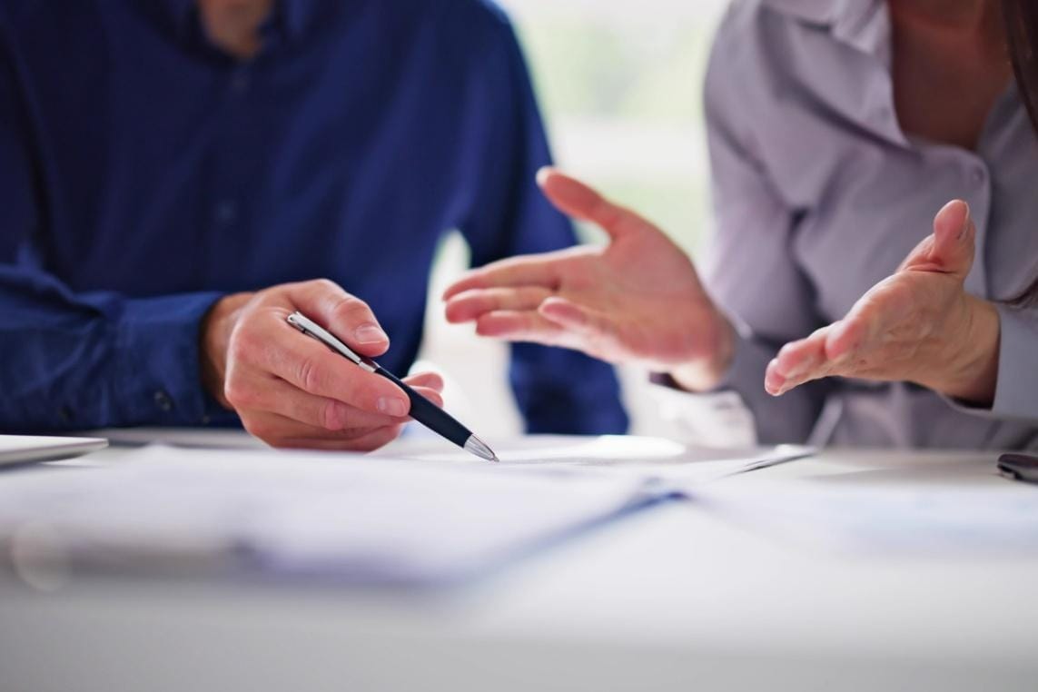 Two people in business attire sitting at a desk, one holding a pen and pointing to documents, the other gesturing with hands during a discussion.