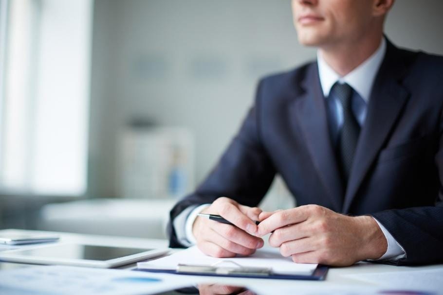A person in a business suit sits at a desk with a tablet, clipboard, and papers, holding a pen and looking away from the camera.