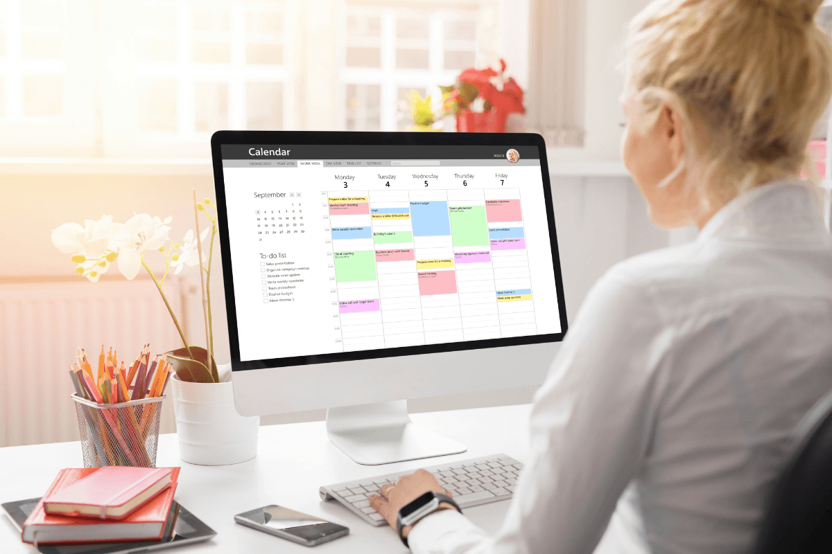 A woman sits at a desk viewing a colorful, detailed weekly calendar on a desktop computer in a bright office setting, planning her week with mindful leadership to enhance mental performance.