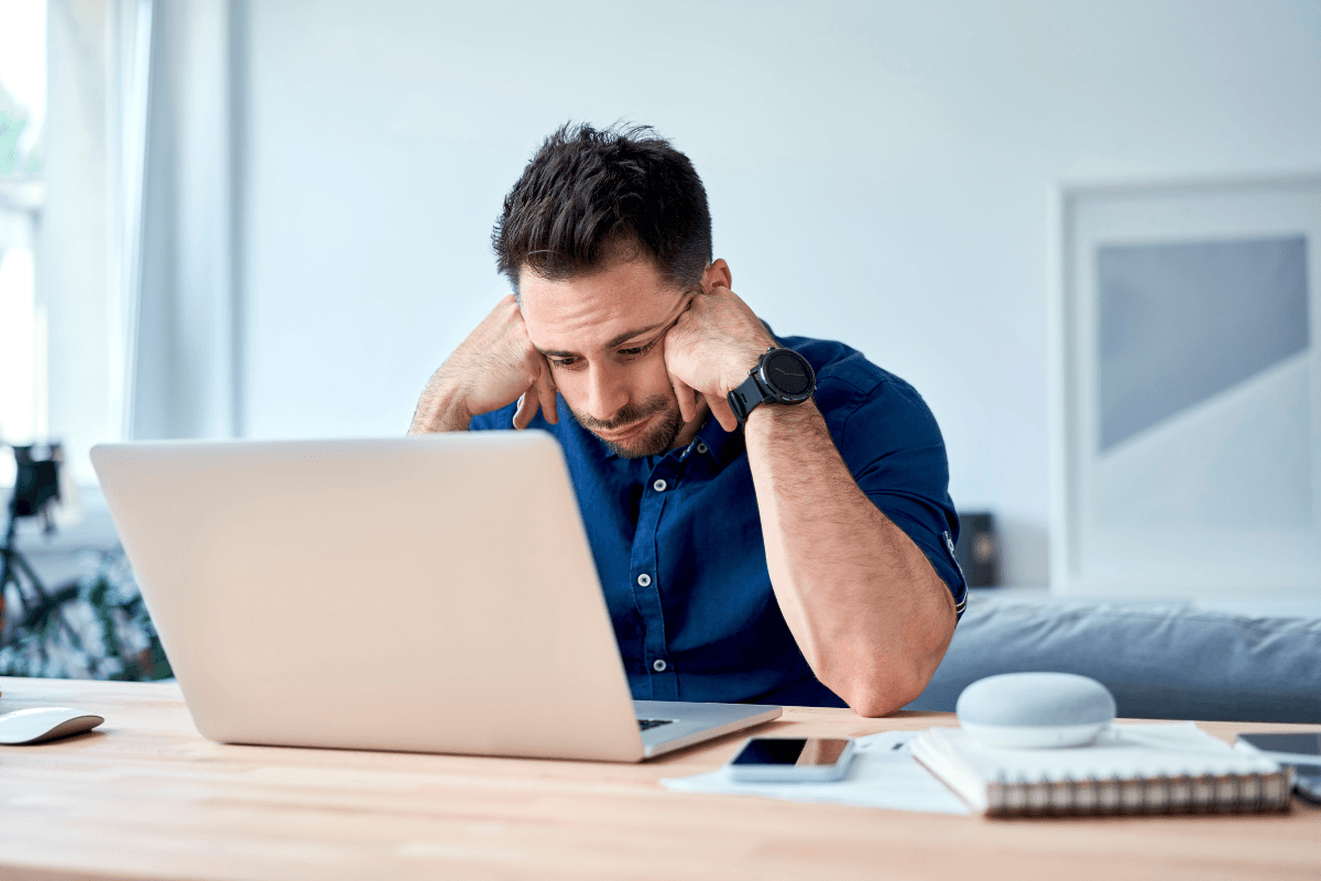 A man sits at a desk looking frustrated while staring at a laptop, with a smartphone, notebook, and smart speaker nearby—highlighting the challenges to mental performance in a fast-paced work environment.