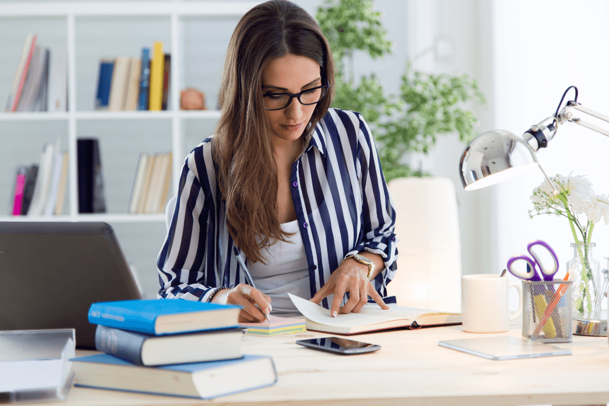 A woman wearing glasses sits at a desk with books, a laptop, and stationery, writing in a notebook under a desk lamp in a home office setting, focused on mindful leadership and boosting her mental performance.