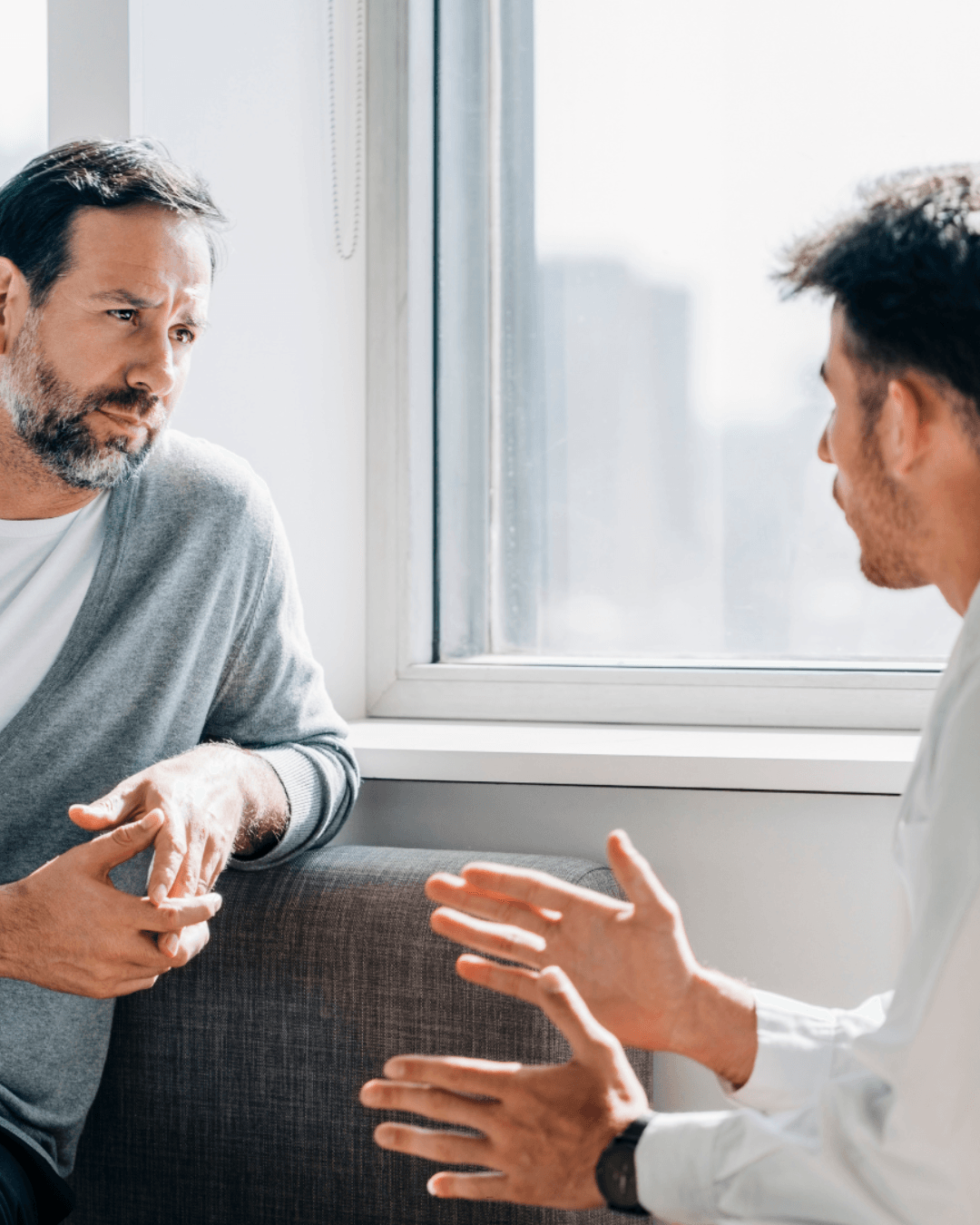 Two men sit near a window having a serious conversation, with one man listening attentively while the other gestures with his hands—an authentic moment of mindful leadership and meaningful connection.