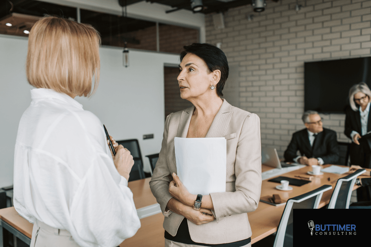 Two women in business attire discuss Mindful Leadership in a conference room, while two colleagues work at a table in the background. A "Buttimer Consulting" logo is visible, highlighting their focus on organizational performance.
