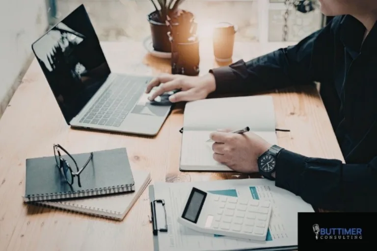 Person working at a desk with a laptop, notepad, calculator, and documents; eyeglasses and a coffee cup sit on the wooden workspace, reflecting mindful leadership and attention to organizational performance.