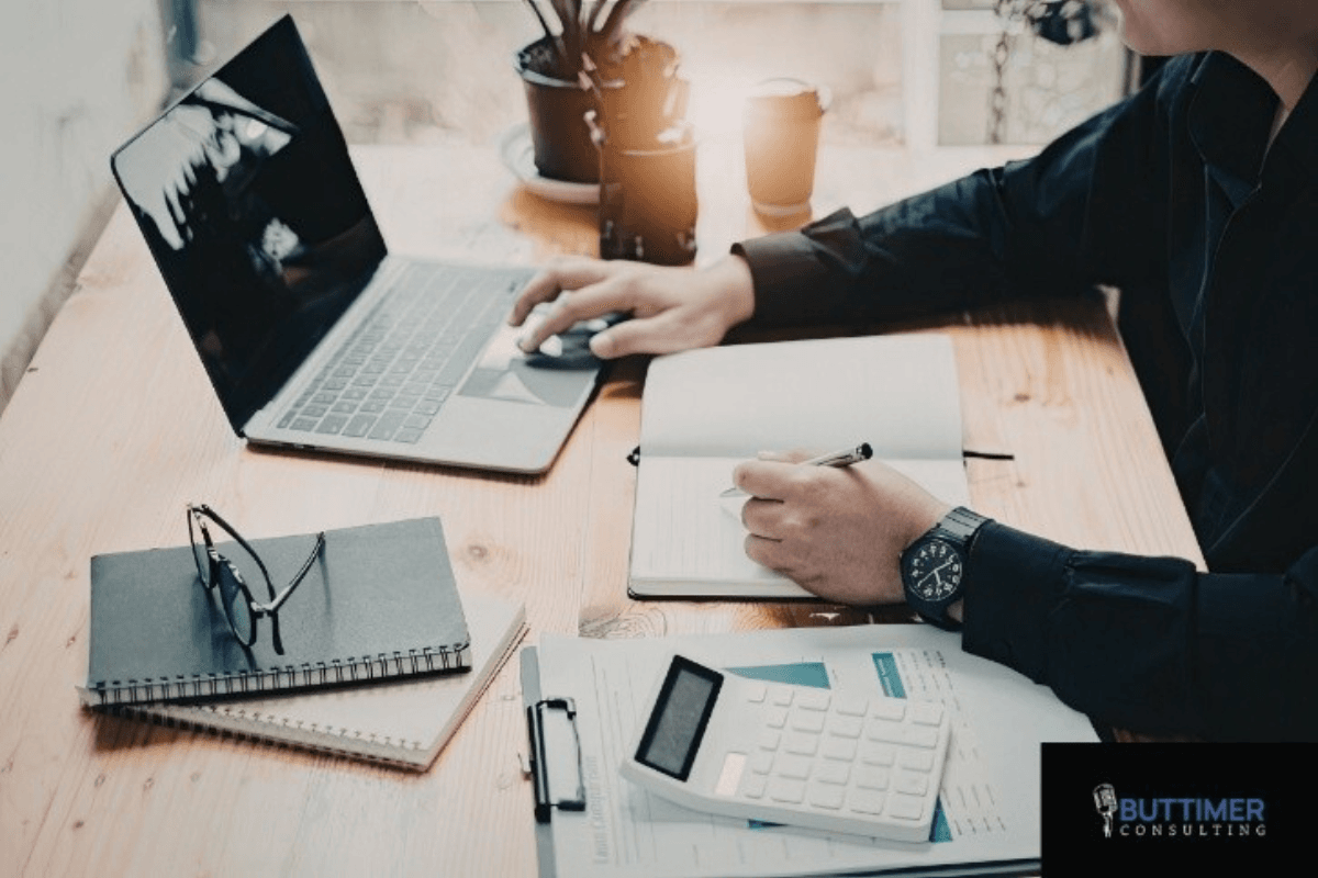 Person working at a desk with a laptop, notepad, calculator, and documents; eyeglasses and a coffee cup sit on the wooden workspace, reflecting mindful leadership and attention to organizational performance.