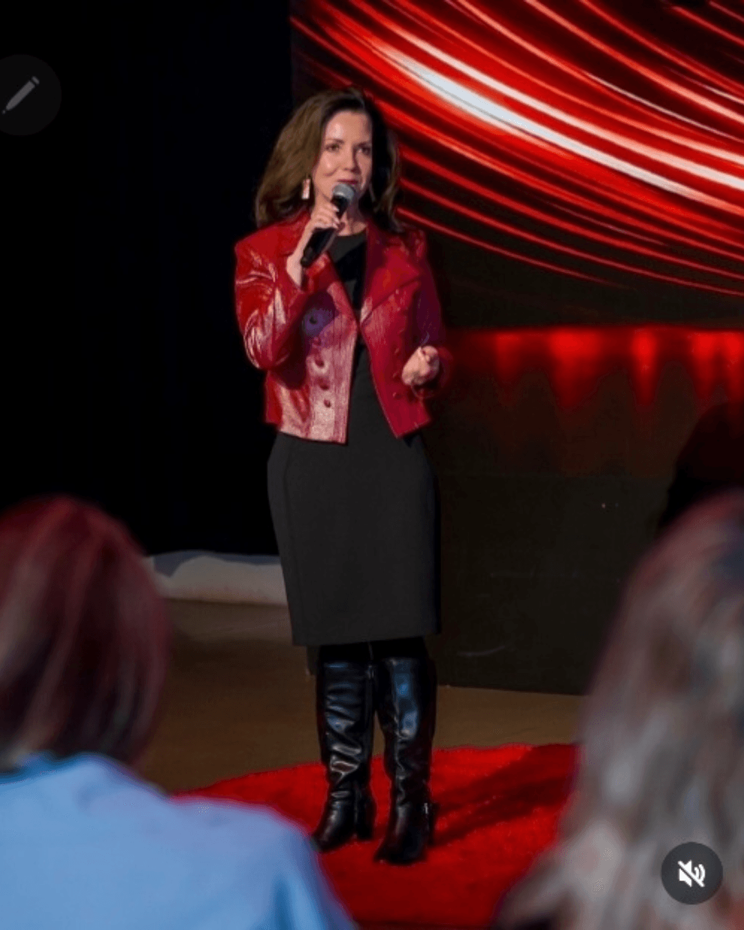 Angela Buttimer, in a red jacket and black dress, stands on stage with a microphone, speaking to an audience as red and black abstract lights glow in the background.
