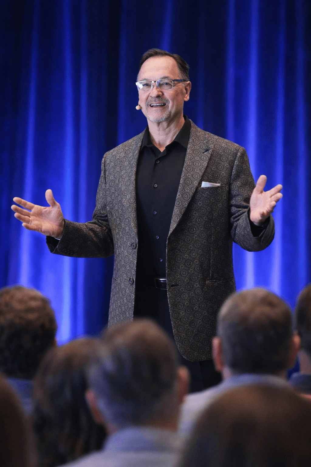 Dennis Buttimer, wearing a patterned blazer and glasses, speaks on stage with a headset microphone in front of an audience, framed by a blue curtain backdrop.