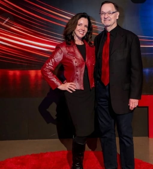 Two adults stand on a red circular carpet in front of a dark backdrop with red light trails. Both wear black outfits with red accents, smiling at the camera, as if sharing something special about the moment.